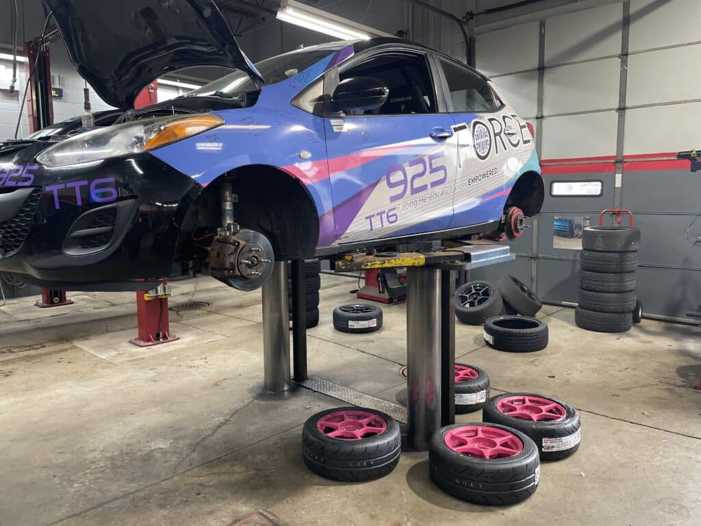 A blue race car on a hydraulic lift inside an auto shop, with its hood open and pink rims stacked nearby for tire work.