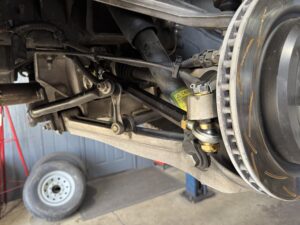 Underbody view of a vehicle showing suspension arms, hoses, wiring, and a removed front wheel area.