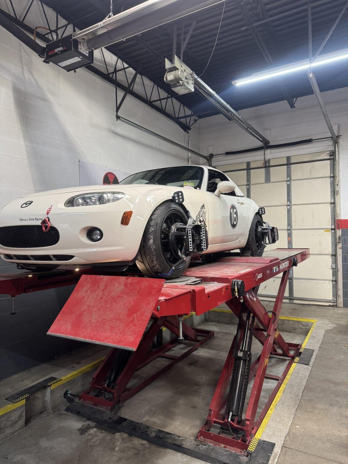White sports car on a red hydraulic lift in a garage, overhead equipment and a closed garage door in the background.