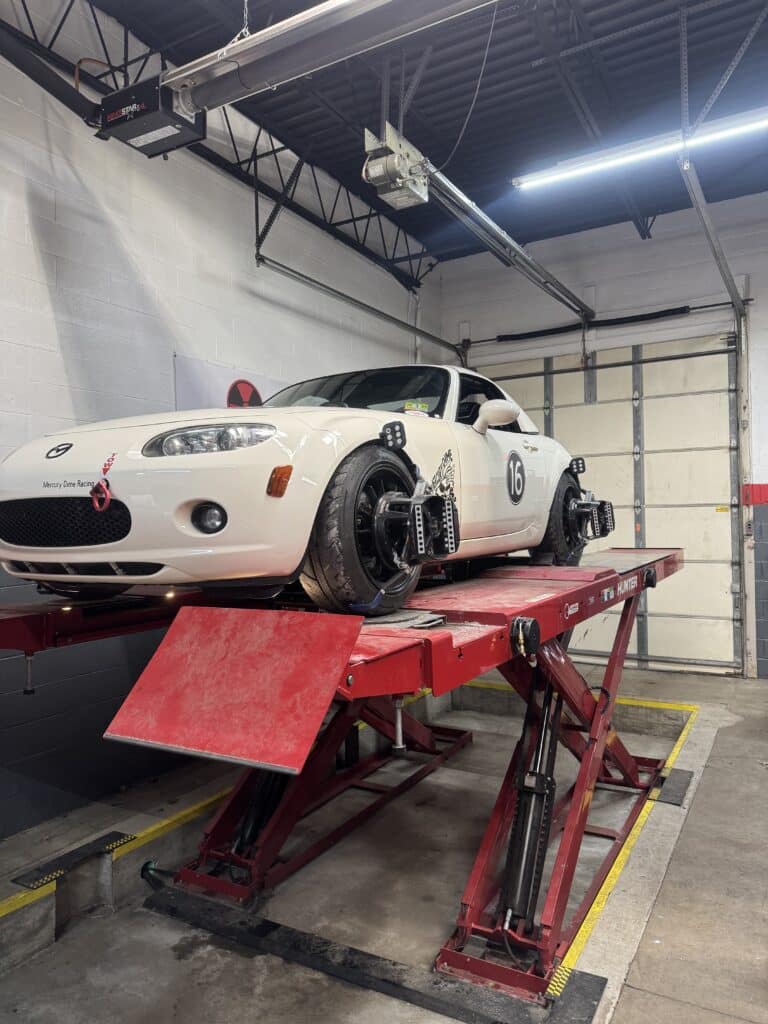 White sports car on a red hydraulic lift in a garage, overhead equipment and a closed garage door in the background.