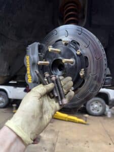 Gloved hand holding multiple brake bolts in front of a slotted brake rotor and AP Racing brake caliper during a repair.