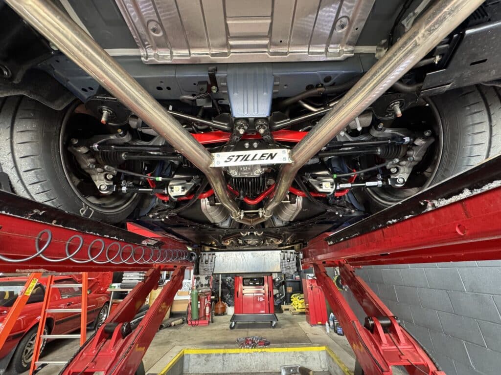 Underside of a car on a hydraulic lift in a repair shop, Stillen plate visible amid the suspension and exhaust components.