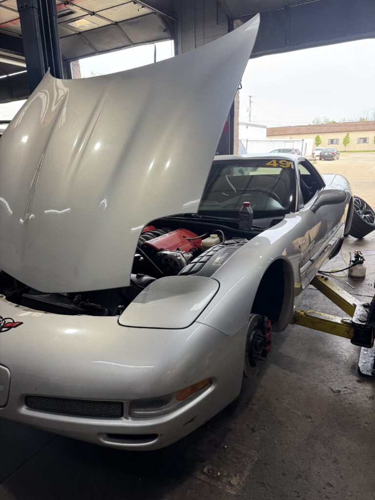 Grey sports car with its hood up in a repair shop, engine visible and a yellow '49' decal on the windshield