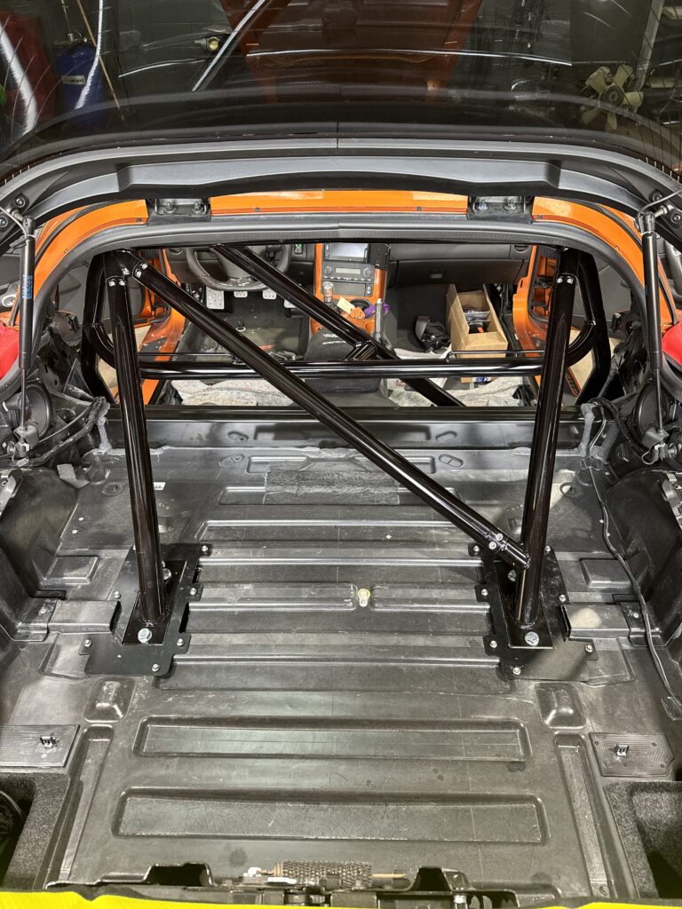 Inside a race car with an orange roll cage and exposed chassis, dashboard and pedals visible behind a rear window bar setup.