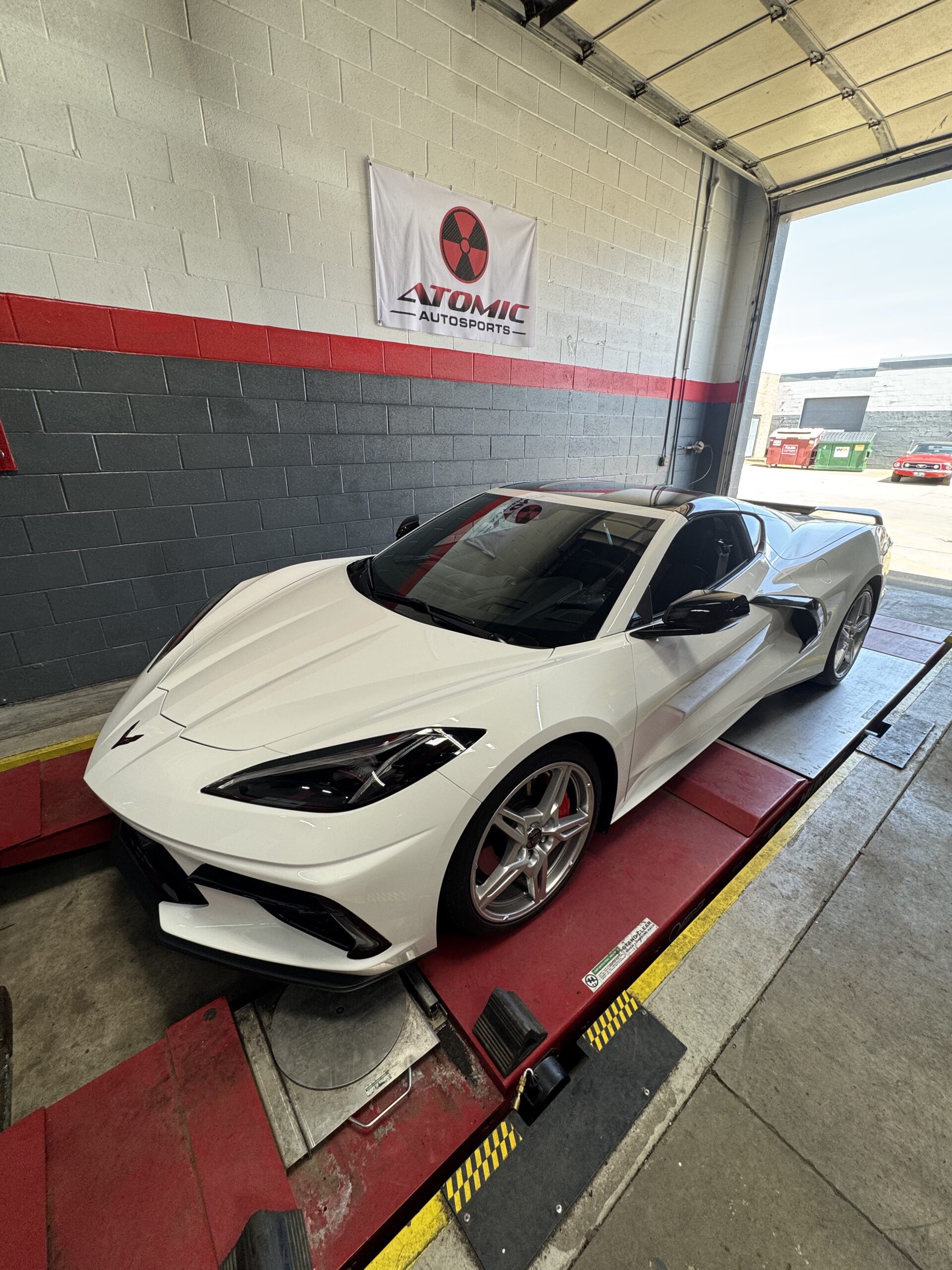 White sports car on a red rolling dyno platform inside a garage, with an Atomic Autosports banner on the wall behind it.