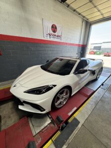 White sports car on a hydraulic lift inside an auto shop, Atomic Autosports banner on the wall behind it.