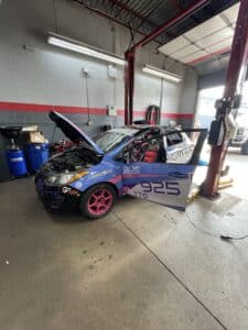 Blue race car with pink rims in a repair shop, hood open and garage equipment nearby (number 925).