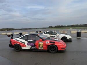 Red and white race car with number 541 and sponsor decals parked on a wet drag strip, with concrete barriers and other cars in the background under a cloudy sky.