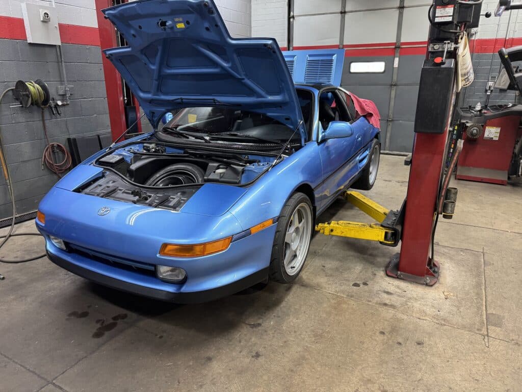 Blue car with its hood raised in a repair shop, engine bay visible and tools in the background.
