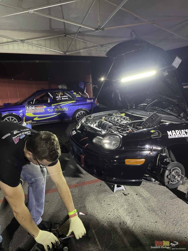 Mechanic wearing gloves crouches by a car engine in a pit area, with a blue Subaru rally car #124 in the background under a canopy