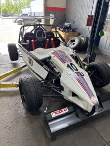 Open-frame race go-kart with red shoulder straps and a white body with red/blue stripes, parked inside a shop with spare tires and tools nearby.