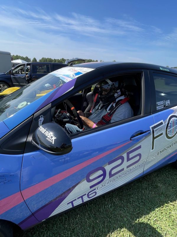 Blue race car with pink accents and a helmeted driver inside, parked on grass at a racing event outside the paddock area