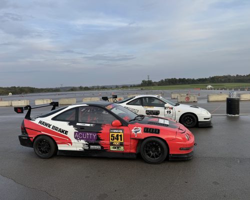 Red and white race car with number 541 and sponsor decals parked on a wet drag strip, with concrete barriers and other cars in the background under a cloudy sky.